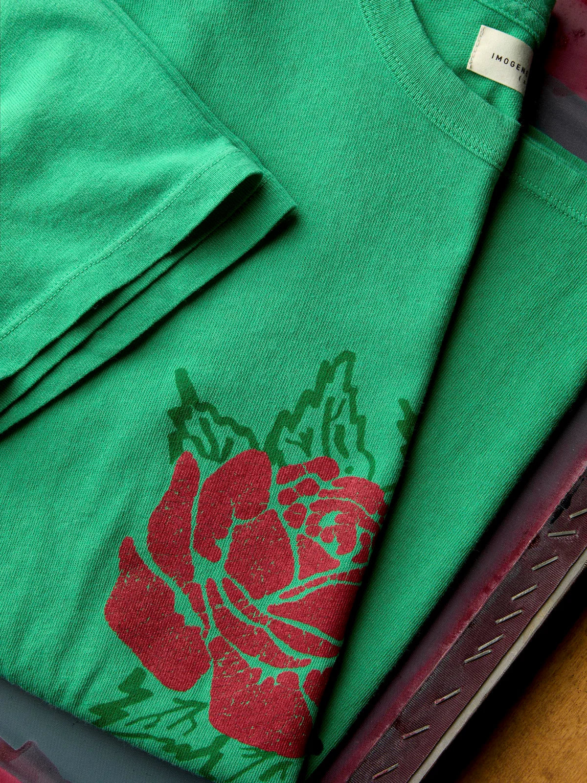 A folded green t-shirt featuring a large red rose graphic with green leaves, displayed on a dark surface with a wooden edge visible.