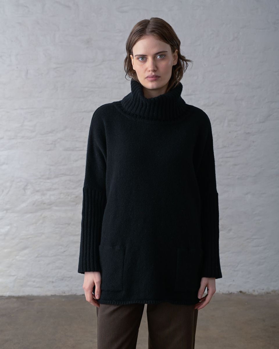 Front view of a woman wearing the BIBICO Adela Jumper in Black, showing the wide ribbed roll neck, batwing sleeves with long ribbed cuffs, two front patch pockets, straight ribbed hem, and oversized longline silhouette in heavyweight black lambswool, styled with brown wide-leg corduroy pants against a whitewashed brick wall
