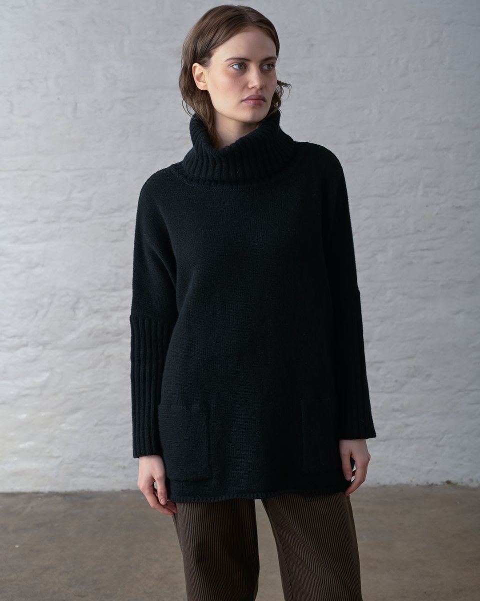 Three-quarter front view of a woman wearing the BIBICO Adela Jumper in Black, showing the wide ribbed roll neck folded, batwing sleeves with long wide ribbed cuffs, front patch pockets, and the oversized longline silhouette in heavyweight black lambswool, styled with brown wide-leg corduroy pants against a whitewashed brick wall
