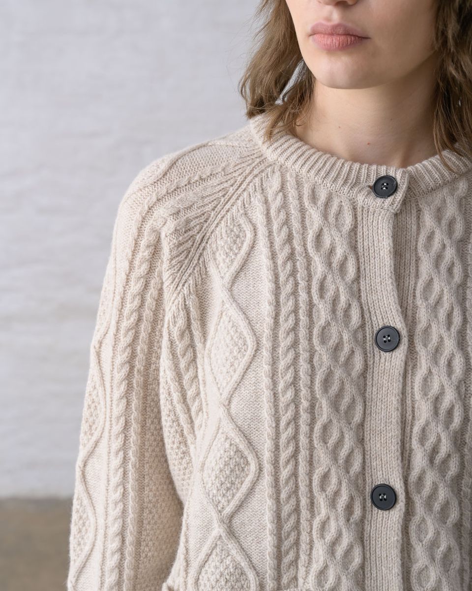 Close detail of the neckline and button front of the BIBICO Derby Aran Cardigan in Oat, showing the ribbed crew neck, imitation corozo buttons, and the intricate diamond and rope Aran cable knit texture of the oat lambswool

