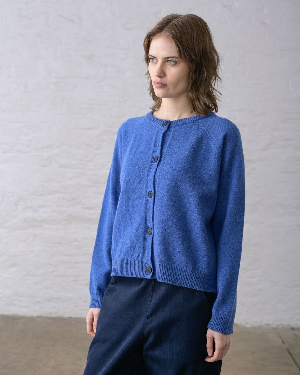 Three-quarter front view of a woman wearing the BIBICO Lottie Cardigan in River buttoned closed, showing the ribbed round neckline, dark imitation corozo button front, raglan sleeves, ribbed hem, and relaxed hip-length silhouette in midweight river blue lambswool, styled with dark navy wide-leg trousers against a whitewashed brick wall
