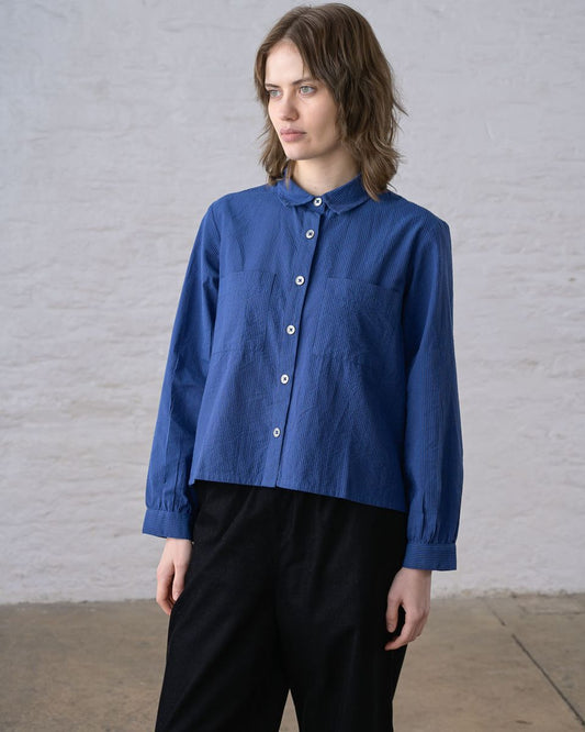 Front view of a woman wearing the BIBICO Ada Cropped Blouse in Blue Petrol Stripe, showing the rounded collar, full button front with imitation shell buttons, two chest patch pockets, and boxy cropped silhouette in fine-woven blue petrol cotton stripe, styled with black wide-leg pants against a whitewashed brick wall
