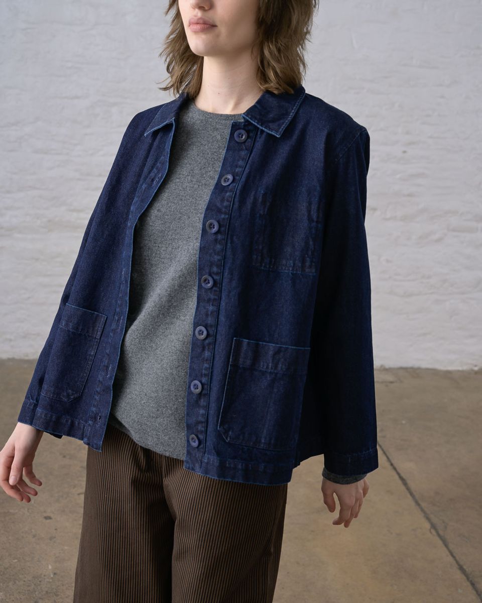 Three-quarter side view of a woman wearing the BIBICO Marcia Jacket in Denim open, showing the pointed collar, button front, hip patch pocket, structured shoulder, and relaxed boxy fit in deep indigo 11.5oz cotton denim, styled with a gray tee and brown wide-leg corduroy pants against a whitewashed brick wall
