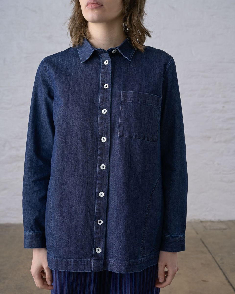 Slightly wider cropped front view of a woman wearing the BIBICO Miriam Tunic in Denim, showing the pointed collar, white button front, chest patch pocket, and relaxed tunic-length silhouette in deep indigo 9oz cotton denim, styled with a blue pleated skirt against a whitewashed brick wall
