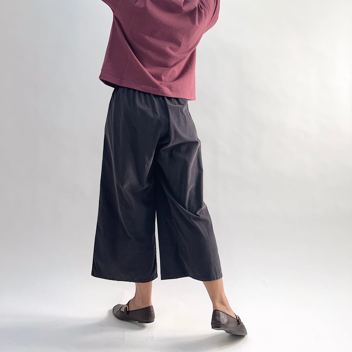 Back view of model wearing the Bryn Walker Corduroy Flood Pants in Fig, showing elastic gathered waistband, wide culotte-style leg, mid-calf flood length, and deep fig pinwheel corduroy, styled with burgundy Double Cotton Tee and brown loafers
