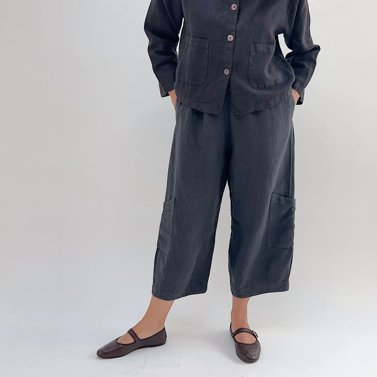 Model wearing the Bryn Walker Heavy Linen Casbah Pant in Moonlight, front view showing fully elastic waistband, two hip pockets, barrel-tapered leg, two large lower-leg patch pockets, cropped ankle length, and deep moonlight heavyweight linen, styled with matching linen jacket and brown Mary Jane flats
