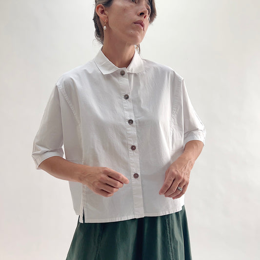 Close front view of model wearing Eleven Stitch chalk camp shirt with hands at hem, showing rounded collar, dark shell buttons, short sleeves with rolled cuffs, slightly cropped fit, side slits, and white color, styled with forest green wide-leg skirt
