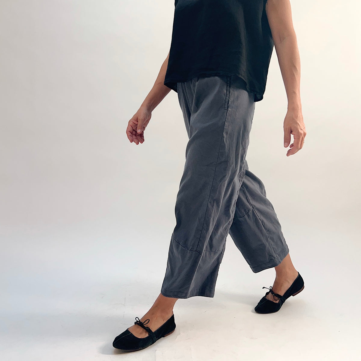 Walking front-angle view of pewter linen-tencel lantern pants showing wide curved leg silhouette in motion, cropped ankle length, paired with a black linen top and black bow-tie ballet flats
