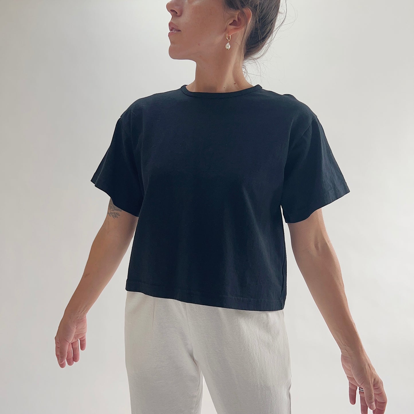 Three-quarter front view of model wearing Eleven Stitch black crop crew tee with arms at sides, showing crew neckline, short sleeves, relaxed boxy fit, slightly cropped length, and black color, styled with white wide-leg pants