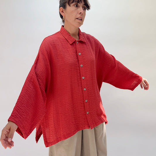 Eleven Stitch Travel Big Shirt in Pimento, three-quarter front view on model in motion, pimento red textured rayon-nylon with point collar, wood button closure, and side slits, paired with khaki wide-leg trousers
