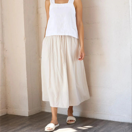Woman wearing a white sleeveless top and beige skirt standing against a plain wall.