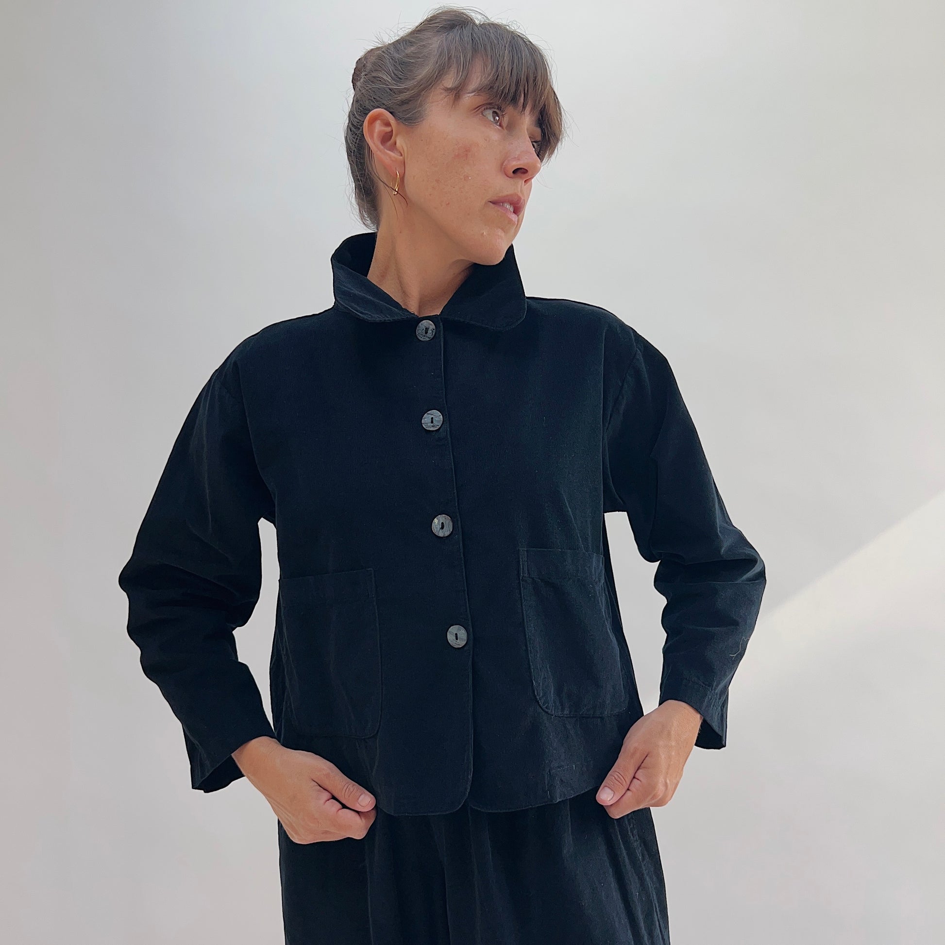 Model with hands in pockets wearing the Bryn Walker Corduroy Audrey Jacket in Black, front view highlighting shell button closure, patch pockets, and pinwheel corduroy texture
