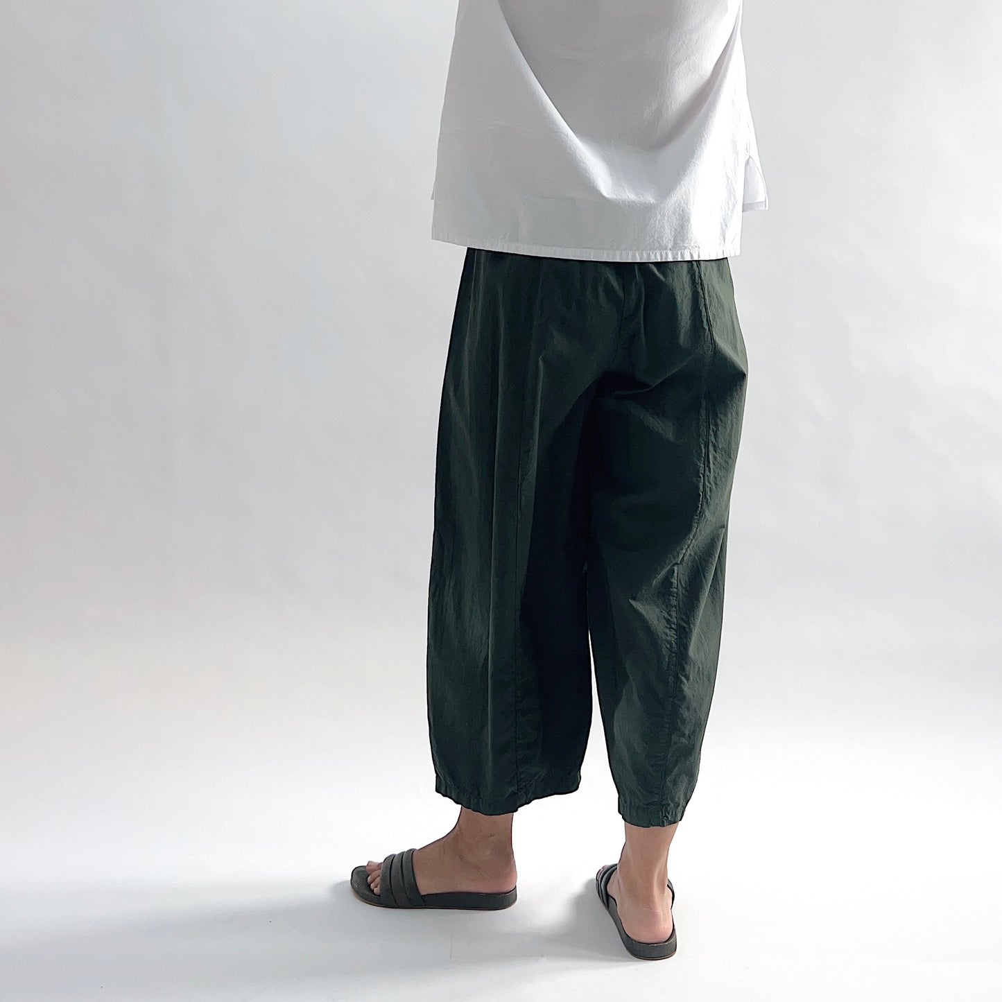 Back view of model wearing Eleven Stitch konbu one pocket pant showing elastic waistband, wide curved-seam cropped leg, large curved side pocket, gathered elastic hem, and deep forest green color, styled with white long sleeve button front shirt and dark slide sandals