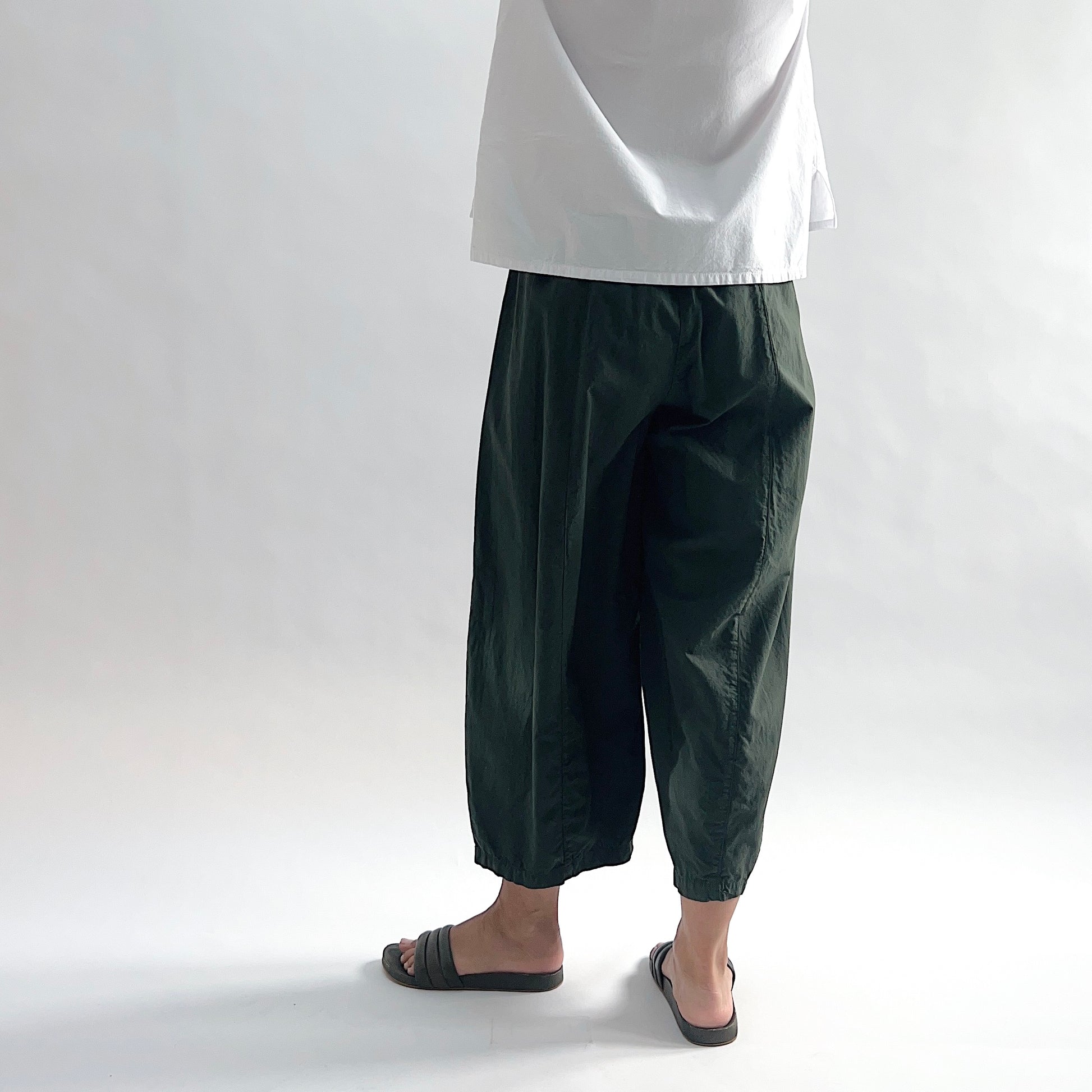 Back view of model wearing Eleven Stitch konbu one pocket pant showing elastic waistband, wide curved-seam cropped leg, large curved side pocket, gathered elastic hem, and deep forest green color, styled with white long sleeve button front shirt and dark slide sandals