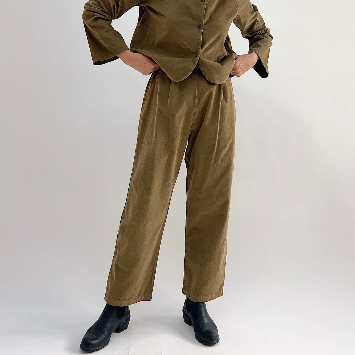 Model with hands on hips wearing the Bryn Walker Corduroy Pleated Pants in Partridge, front view showing front pleats, straight leg silhouette, and olive pinwheel corduroy, paired with matching Standing Collar Crop Jacket and black Chelsea boots
