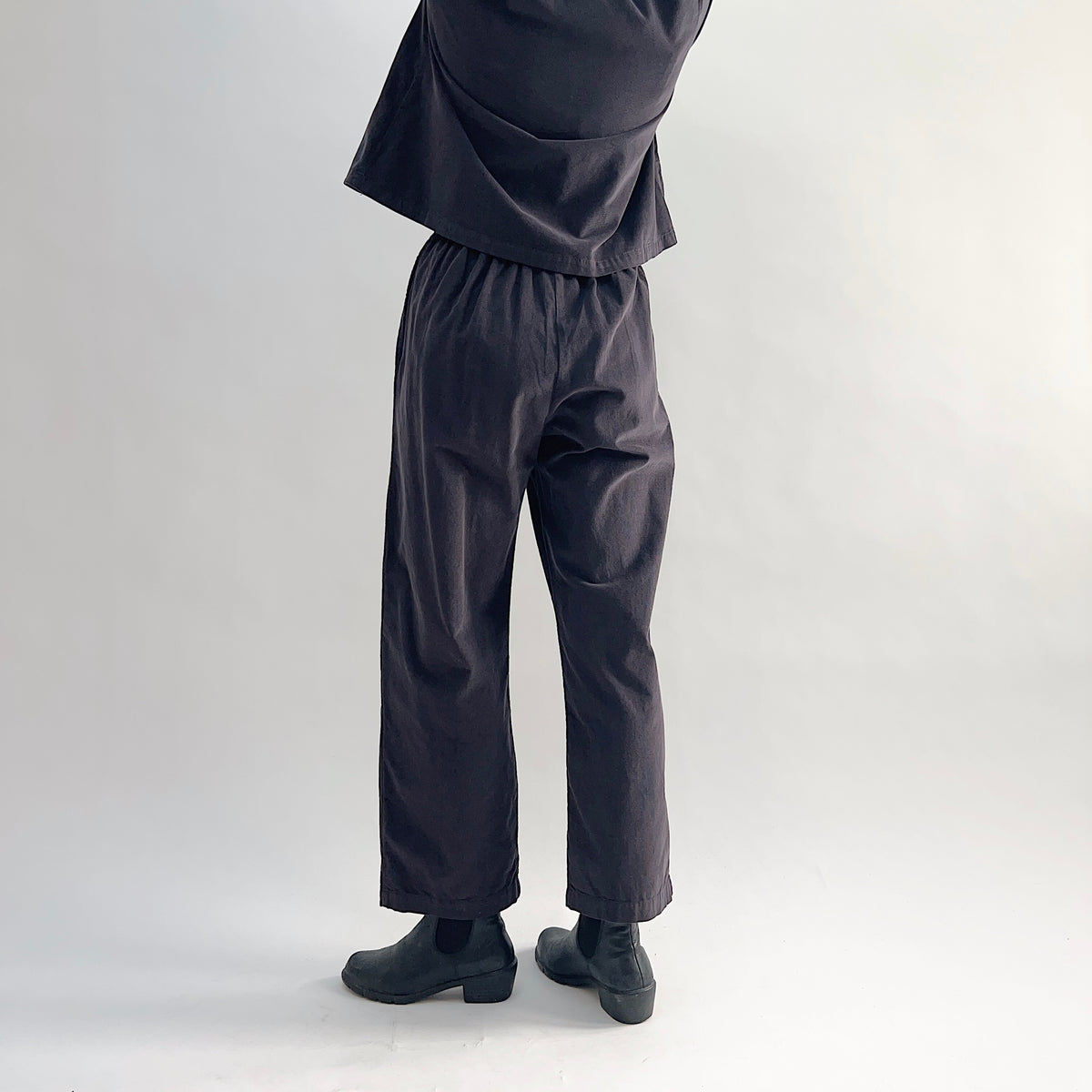 Back view of model wearing the Bryn Walker Corduroy Pleated Pants in Fig, showing elastic back waistband, tapered straight leg silhouette, and deep fig pinwheel corduroy drape, styled with matching Standing Collar Crop Jacket and black Chelsea boots
