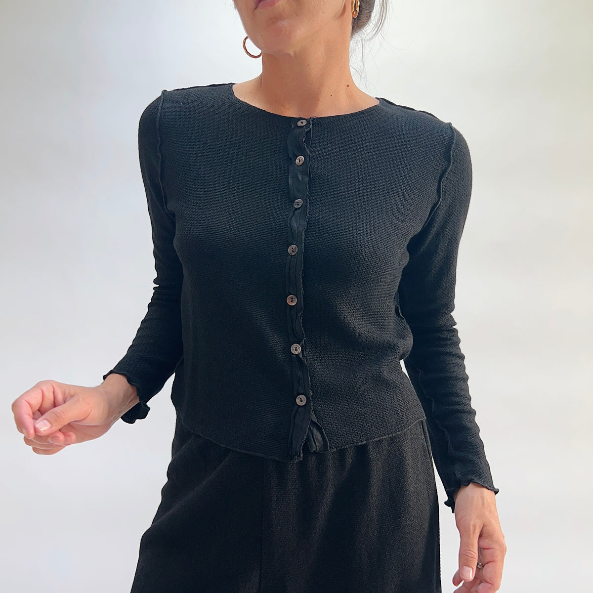 Model wearing the Cut Loose Marled Knit Cropped Cardigan in Black, three-quarter front view, showing round neckline, scalloped lettuce edges, full button front with dark shell buttons, and cropped straight hem of black marled knit cardigan, styled with a black skirt
