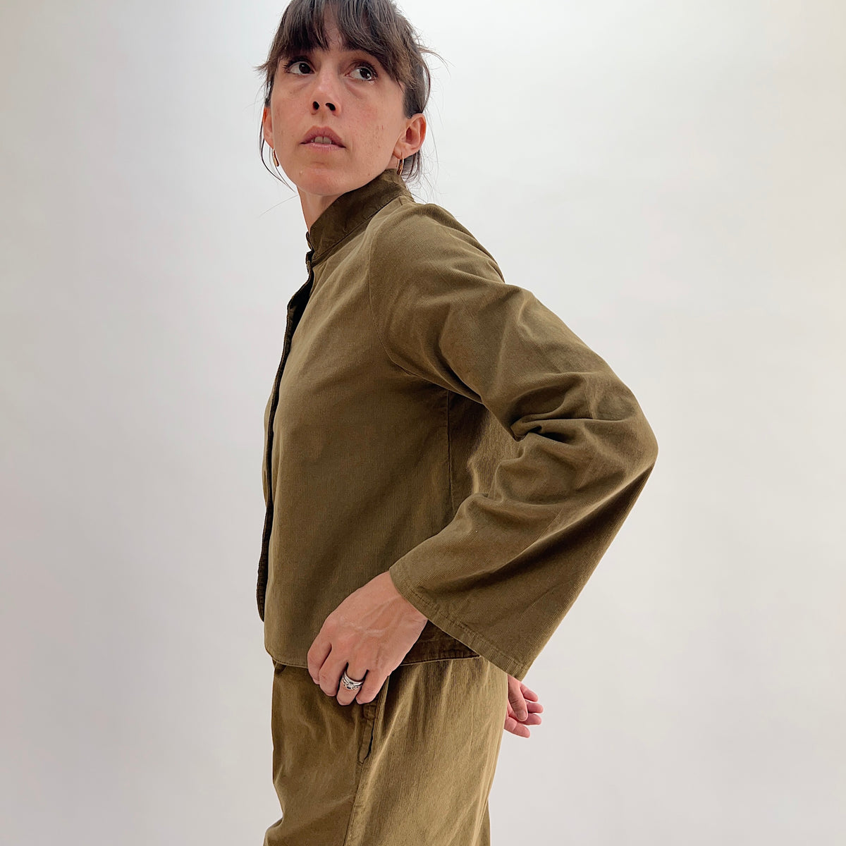 Side profile of model wearing the Bryn Walker Corduroy Standing Collar Crop Jacket in Partridge, highlighting ruffled standing collar, wide sleeve, cropped hem, and olive pinwheel corduroy drape, paired with matching pants
