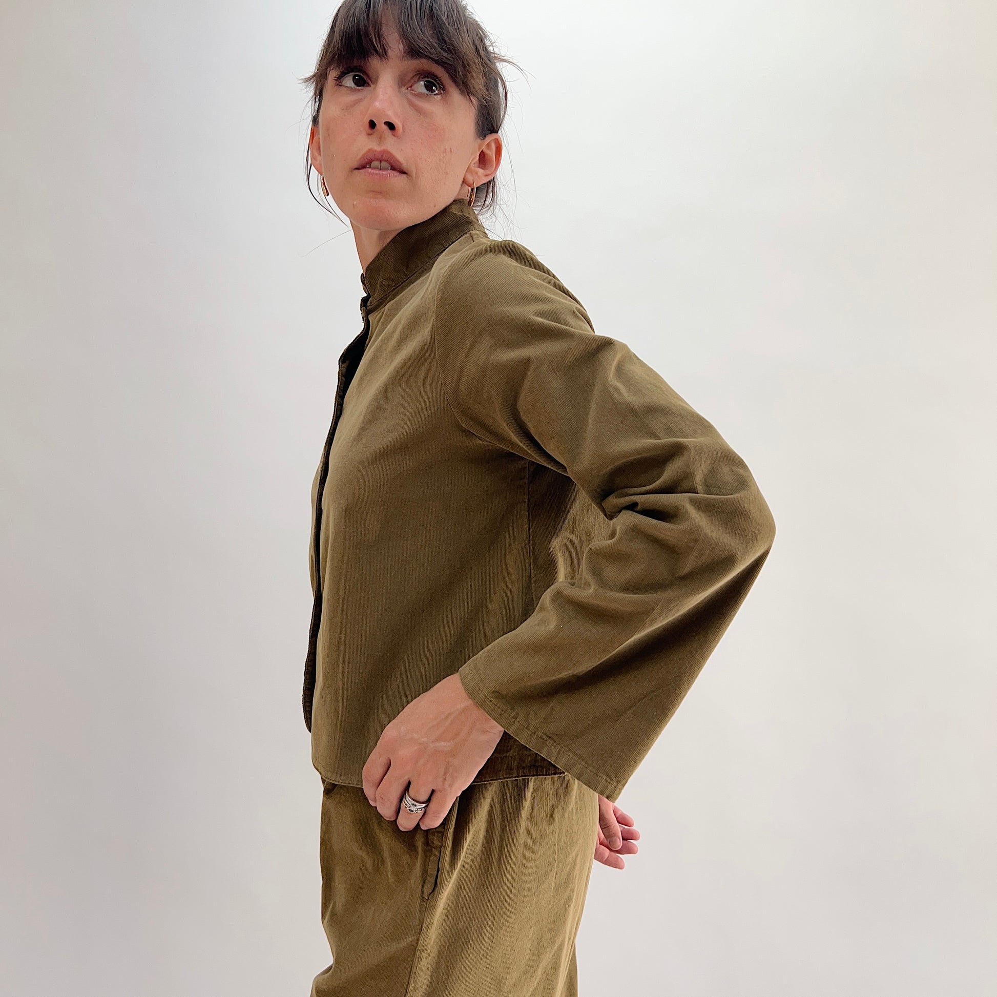 Side profile of model wearing the Bryn Walker Corduroy Standing Collar Crop Jacket in Partridge, highlighting ruffled standing collar, wide sleeve, cropped hem, and olive pinwheel corduroy drape, paired with matching pants
