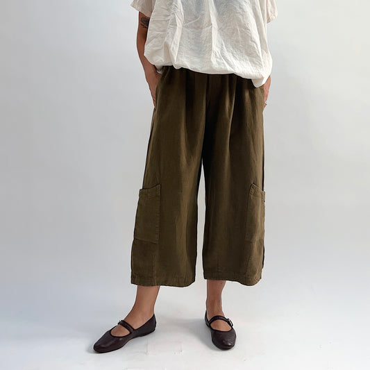 Model wearing the Bryn Walker Heavy Linen Casbah Pant in Partridge, full front view highlighting elastic waistband, two patch pockets, cropped wide-leg hem, and olive linen texture, with dark Mary Jane flats
