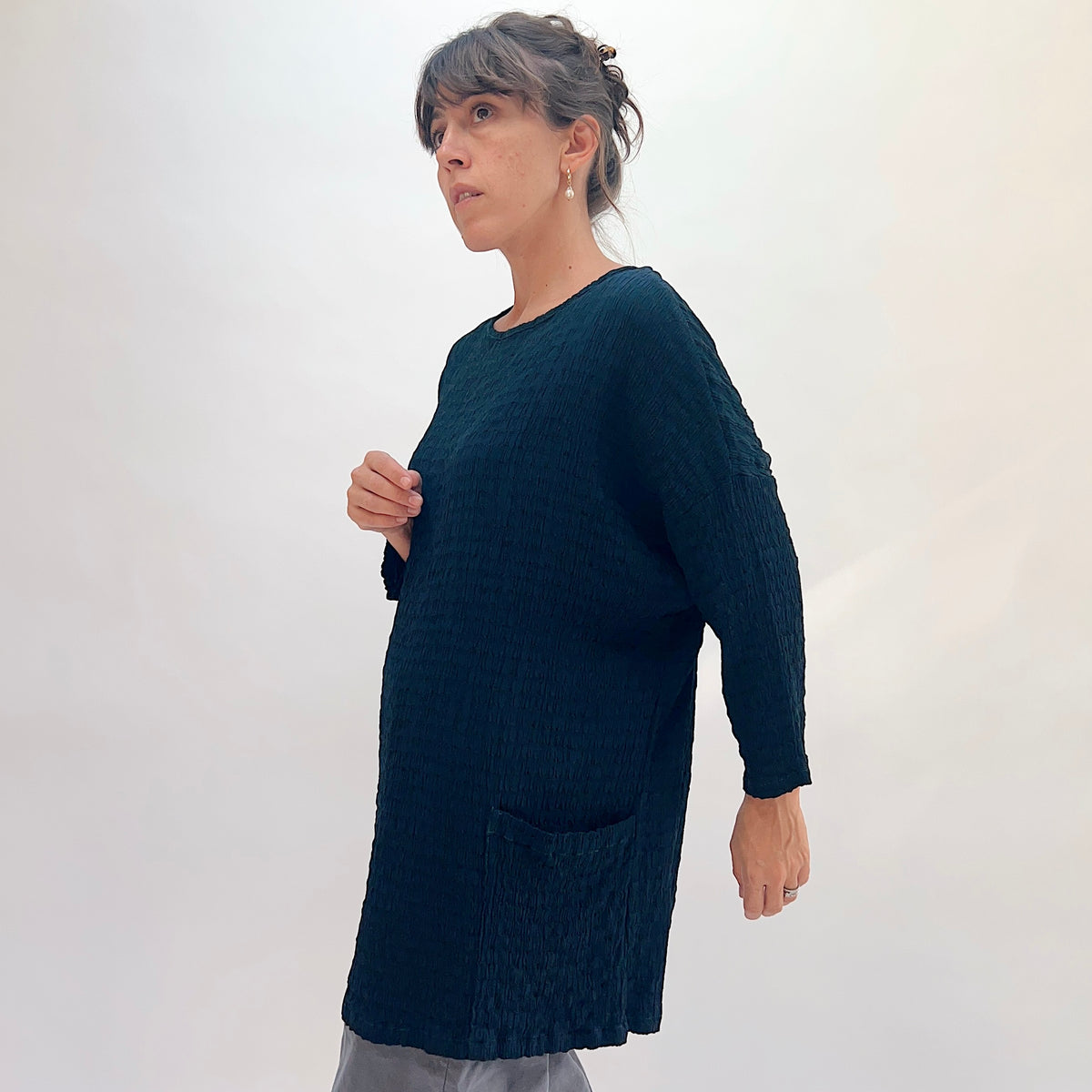 Side view of teal crinkle-texture one-size pocket tunic showing scoop neckline, 3/4-length sleeves, and front patch pocket, paired with grey wide-leg pants, pearl drop earrings
