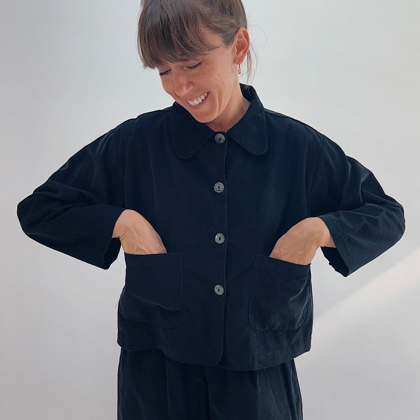 Model smiling with hands in patch pockets of the Bryn Walker Corduroy Audrey Jacket in Black, front view showing rounded collar and shell buttons
