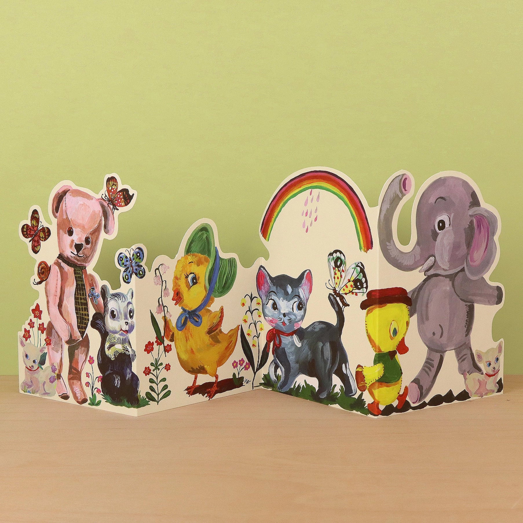Plushie Party die-cut concertina card by Nathalie Lété, partially open showing a pink teddy bear with a tie and a skunk alongside a black cat, yellow chick, and elephant, with butterflies and a rainbow, against a green background