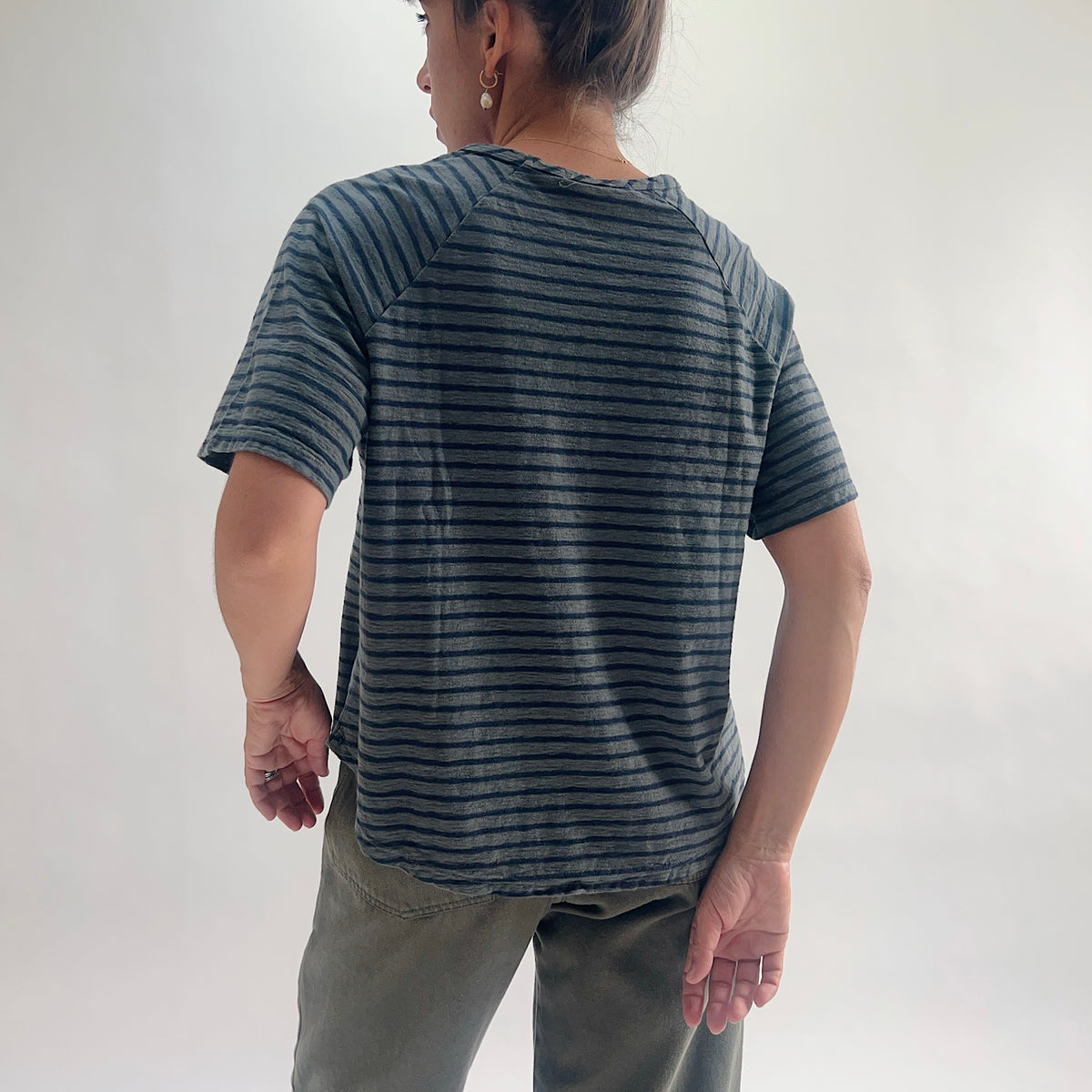 Back view of model wearing an anthracite and navy striped boxy raglan tee with short sleeves and a curved hem, styled with olive green pants against a white background.
