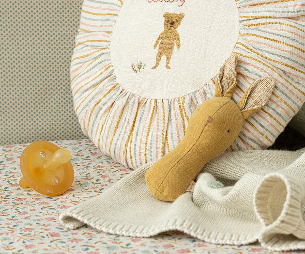 Maileg Lullaby Friends Bunny Rattle in Dusty Yellow styled in a nursery scene with a striped sunburst pillow, cream knit blanket, and amber rubber bunny — a warm golden-yellow linen bunny rattle with upright ears
