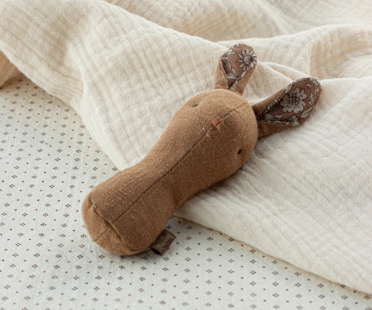 Maileg Lullaby Friends Bunny Rattle in Chocolate resting against a soft cream muslin blanket — a warm brown linen bunny rattle with floral-lined ears
