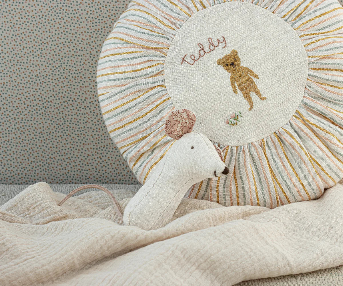 Maileg Lullaby Friends Mouse Rattle in Natural resting against a striped sunburst nursery pillow with a cream muslin blanket — a natural linen mouse rattle with floral-lined ears and pink tail
