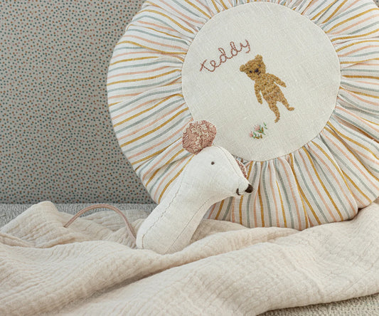 Maileg Lullaby Friends Mouse Rattle in Natural resting against a striped sunburst nursery pillow with a cream muslin blanket — a natural linen mouse rattle with floral-lined ears and pink tail
