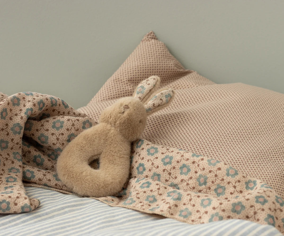 Maileg Rabbit Rattle in Peachy Cream nestled in a floral muslin blanket on a bed with a waffle pillow — a plush peachy-cream bunny ring rattle with floral-lined ears
