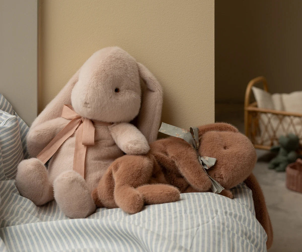 Maileg Small Sleeping Plush Bunny in Soft Brown styled in a nursery scene — a soft brown sleeping bunny with a blue floral bow nestled alongside a larger blush pink Maileg bunny on a striped linen cushion, with a rattan basket in the background

