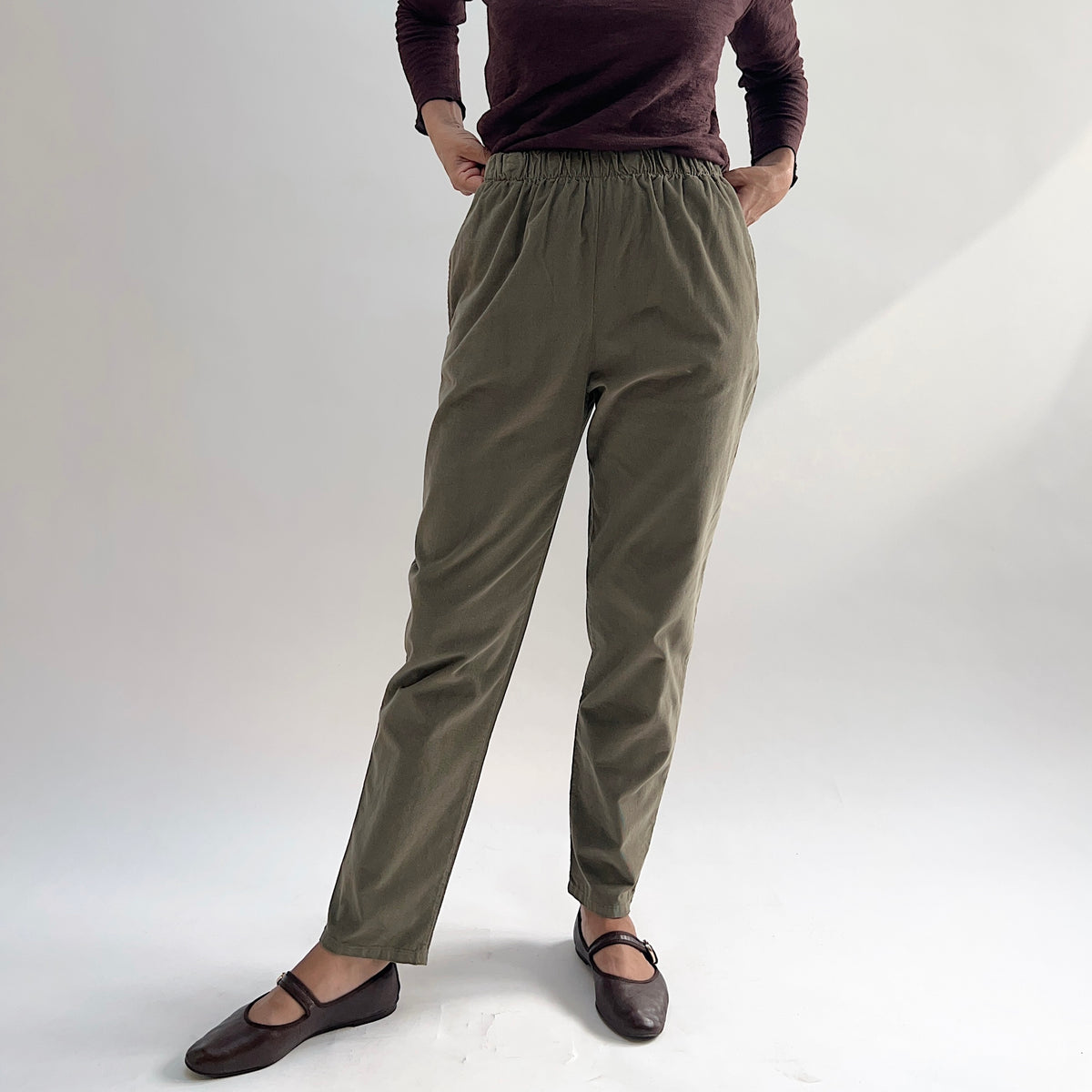 Model with hands in pockets wearing the Bryn Walker Corduroy Boslow Pants in Jasper, full front view highlighting fully elastic waistband, side pockets, relaxed tapered silhouette, and olive jasper pinwheel corduroy, styled with burgundy Double Cotton Tee and brown Mary Jane flats
