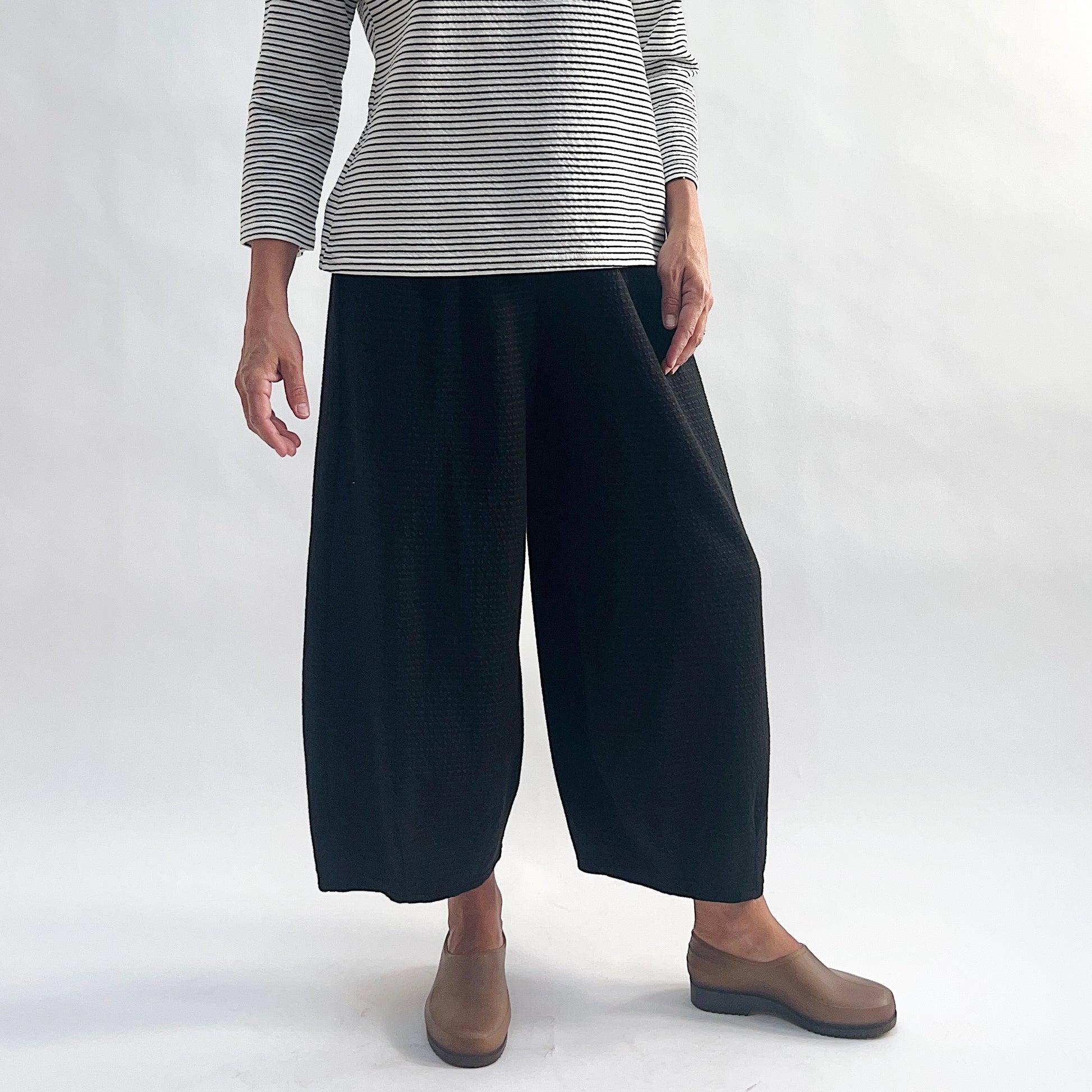 Front view of model wearing black crinkle-texture cropped wide-leg pants with elastic waist, arms at sides, showing the relaxed rounded silhouette and tapered ankle, styled with a white and black stripe long-sleeve top and taupe slip-on shoes against a white background.
