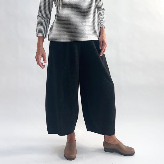 Front view of model wearing black crinkle-texture cropped wide-leg pants with elastic waist, arms at sides, showing the relaxed rounded silhouette and tapered ankle, styled with a white and black stripe long-sleeve top and taupe slip-on shoes against a white background.
