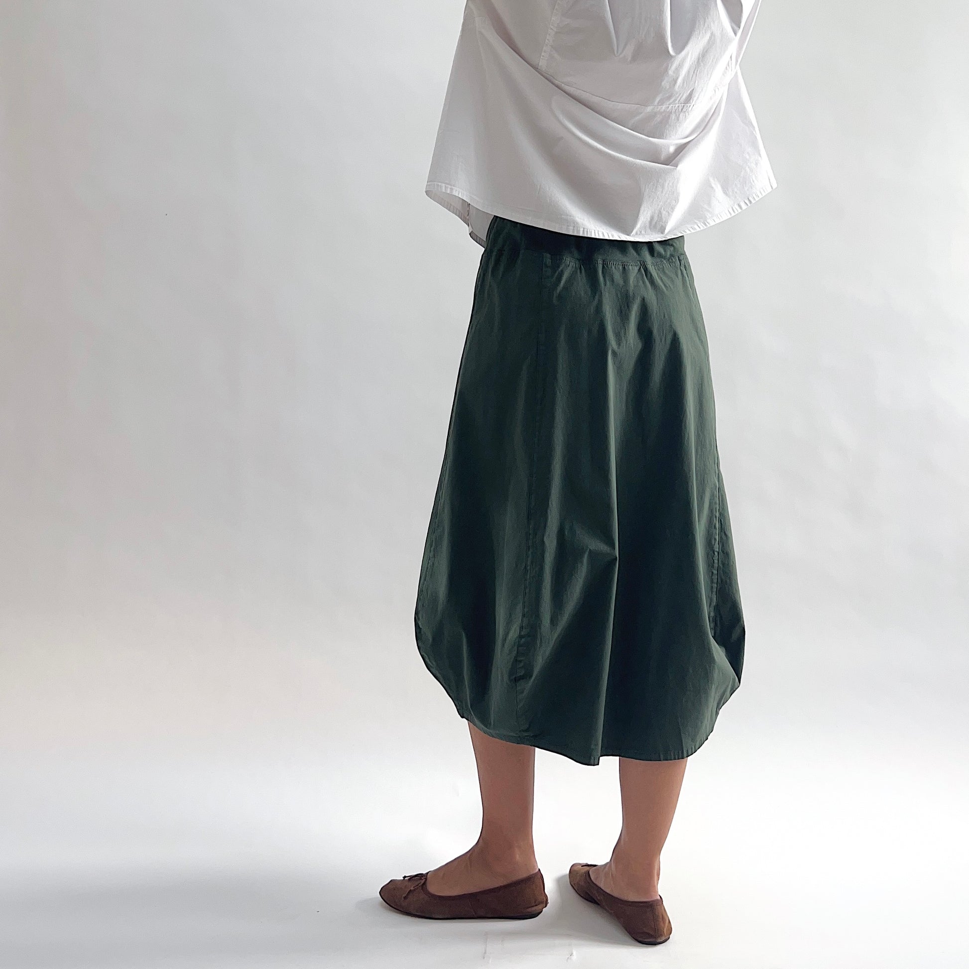 Three-quarter back view of model wearing Eleven Stitch konbu rib yoke skirt, showing elastic knit waistband, curved seam paneling, sculptural bubble hem, below-the-knee length, and deep forest green color, styled with white chalk camp shirt and brown ballet flats