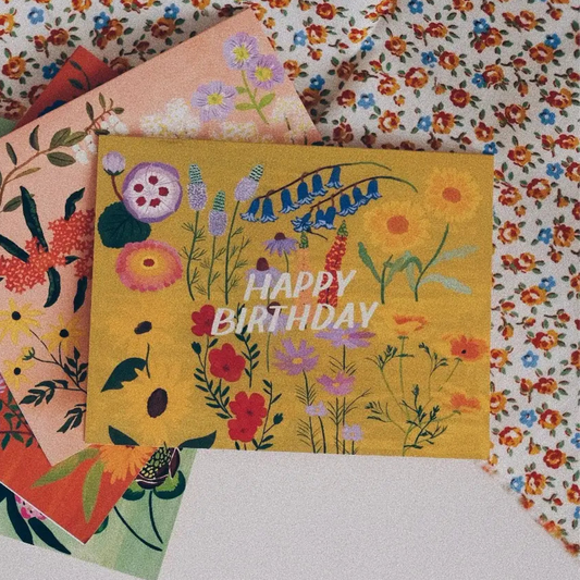 Small Adventure Yellow Floral Birthday Card, colorful illustrated flowers on a mustard yellow background with white “Happy Birthday” lettering.
