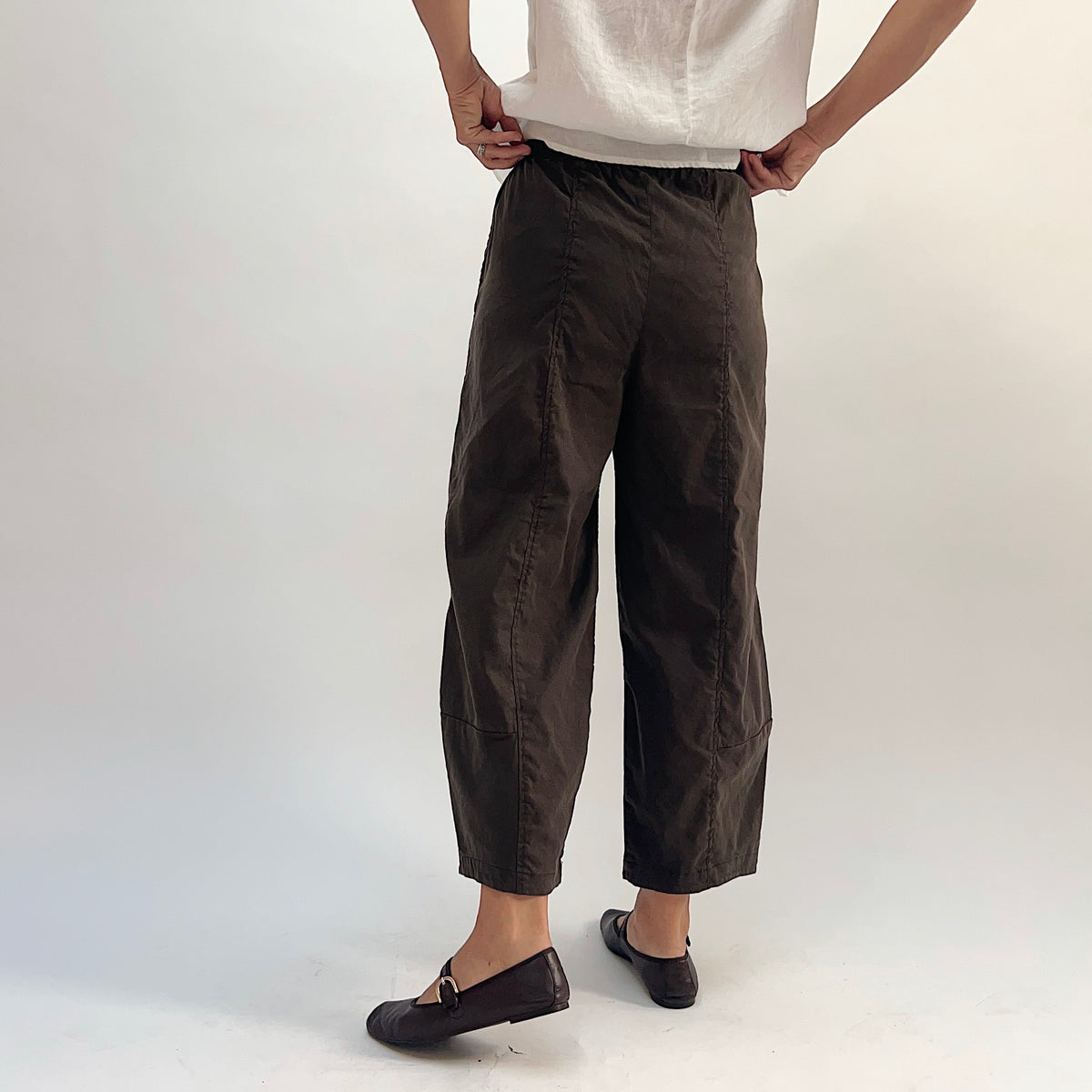 Back view of walnut linen-tencel lantern pants showing curved seam detail down both legs, elastic waistband, and cropped ankle length, worn with a white linen top and black loafers, hands on hips
