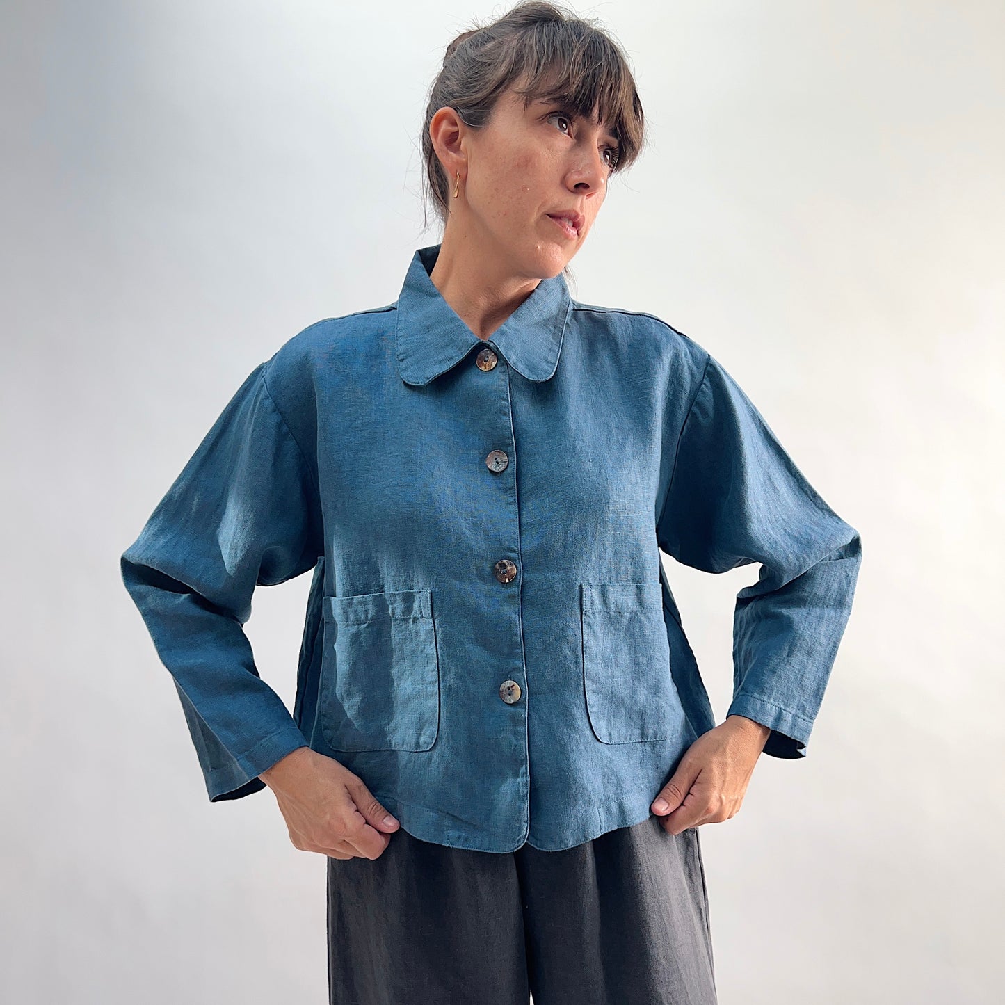 Model wearing the Bryn Walker Heavy Linen Audrey Jacket in Vitznau, front view with hands on hips, showing rounded collar, shell buttons, and patch pockets, paired with dark trousers
