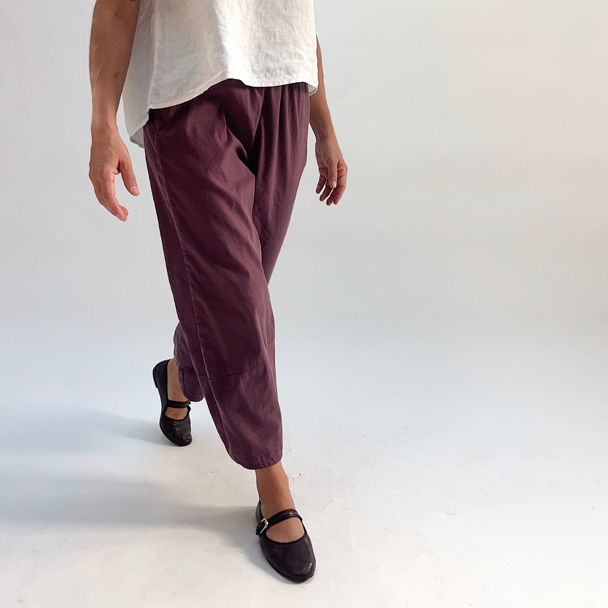 Model walking in russet linen-tencel lantern pants with curved seam legs and elastic waistband, paired with a white top and dark Mary Jane flats, side-angle view
