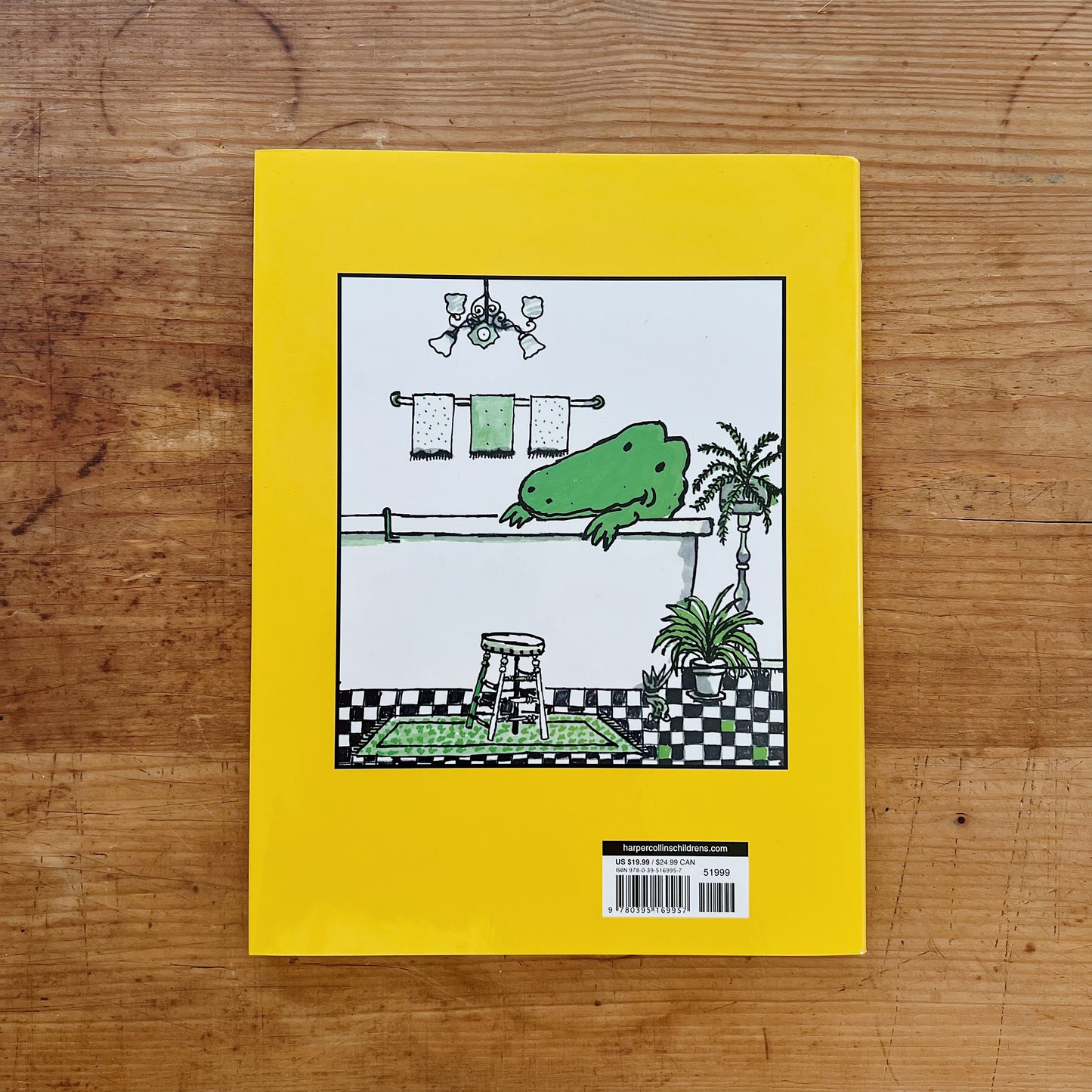 Back cover of Lyle, Lyle, Crocodile hardcover displayed flat on a warm wood surface, featuring a solid bright yellow background. A centered rectangular vignette illustration shows Lyle the crocodile — green and spotted — draped over the edge of a white claw-foot bathtub in a black-and-white checkerboard bathroom, with hanging towels, a wooden stool, and a potted fern. Below the vignette, a white label reads harpercollinschildrens.com and a barcode with retail price of $19.99 USD and $24.99 CAN.
