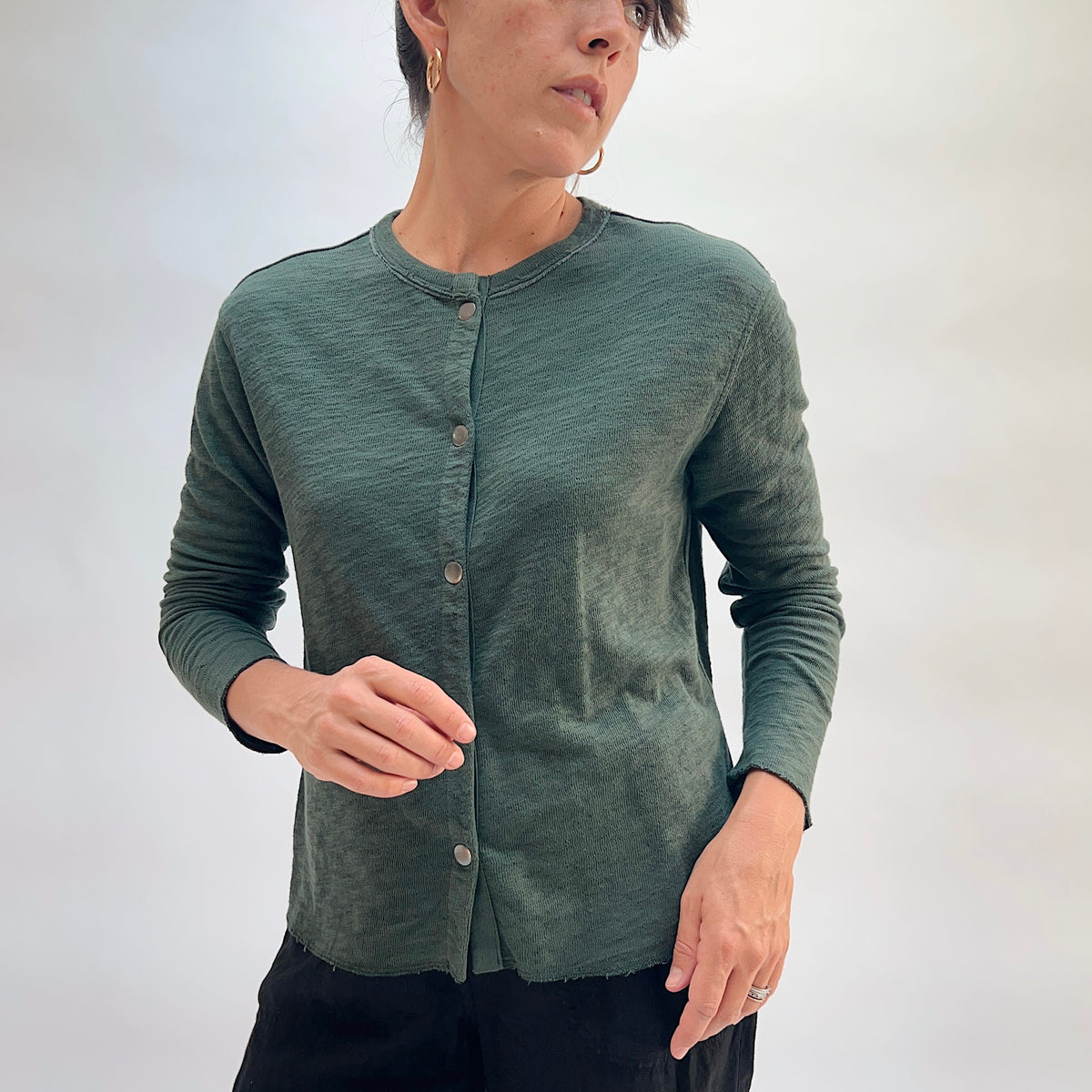 Model wearing the Cut Loose Snap Front Cardigan in Konbu, front view, fitted snap-front cotton cardigan in deep teal green with raw hem, styled with black trousers
