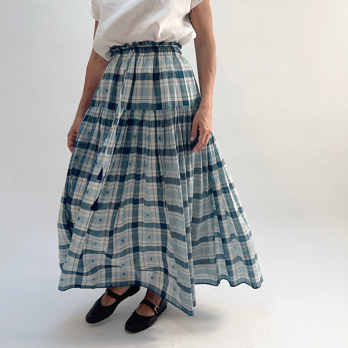 DVAA Denise Skirt in Blue Plaid, tiered cotton maxi skirt with tassel tie at the waist, worn with a white tee and black Mary Jane flats — angled front view showing movement

