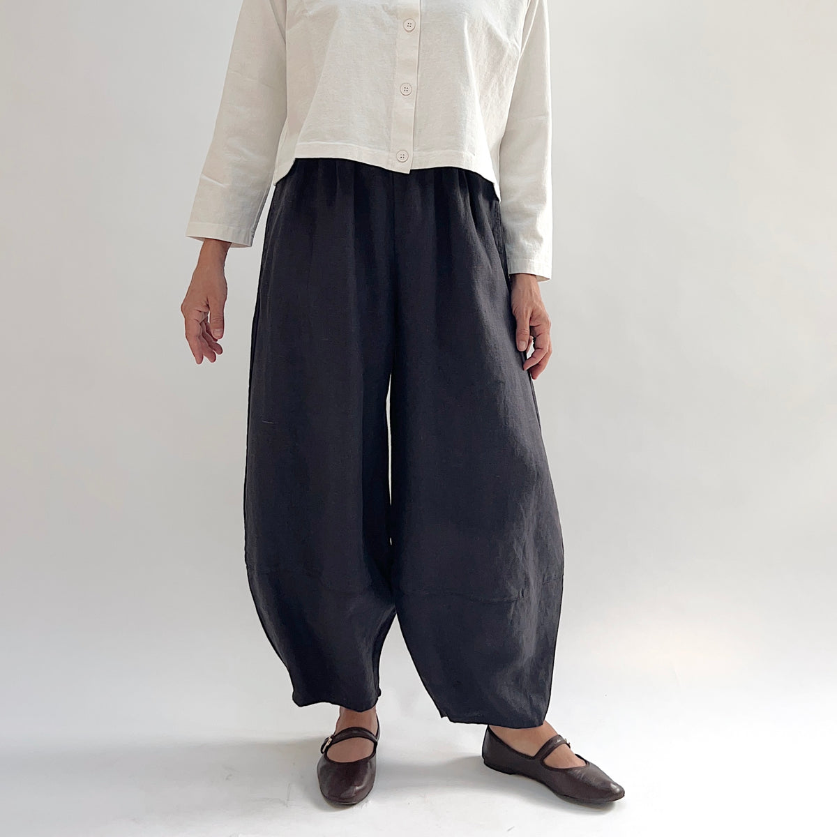 Model wearing the Bryn Walker Heavy Linen Oliver Pant in Fig, full front view highlighting barrel-leg cocoon silhouette, elastic waistband, and tapered ankle in deep fig heavy linen, styled with cream button-up and brown Mary Jane flats
