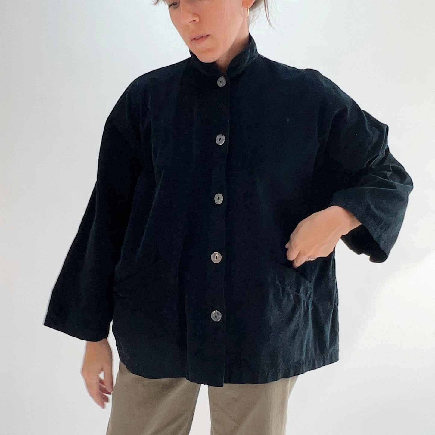Model wearing the Bryn Walker Corduroy Danuta Jacket in Black, cropped front view showing standing collar, shell button closure, wide dolman sleeves, front patch pocket, and black pinwheel corduroy, styled with olive pants
