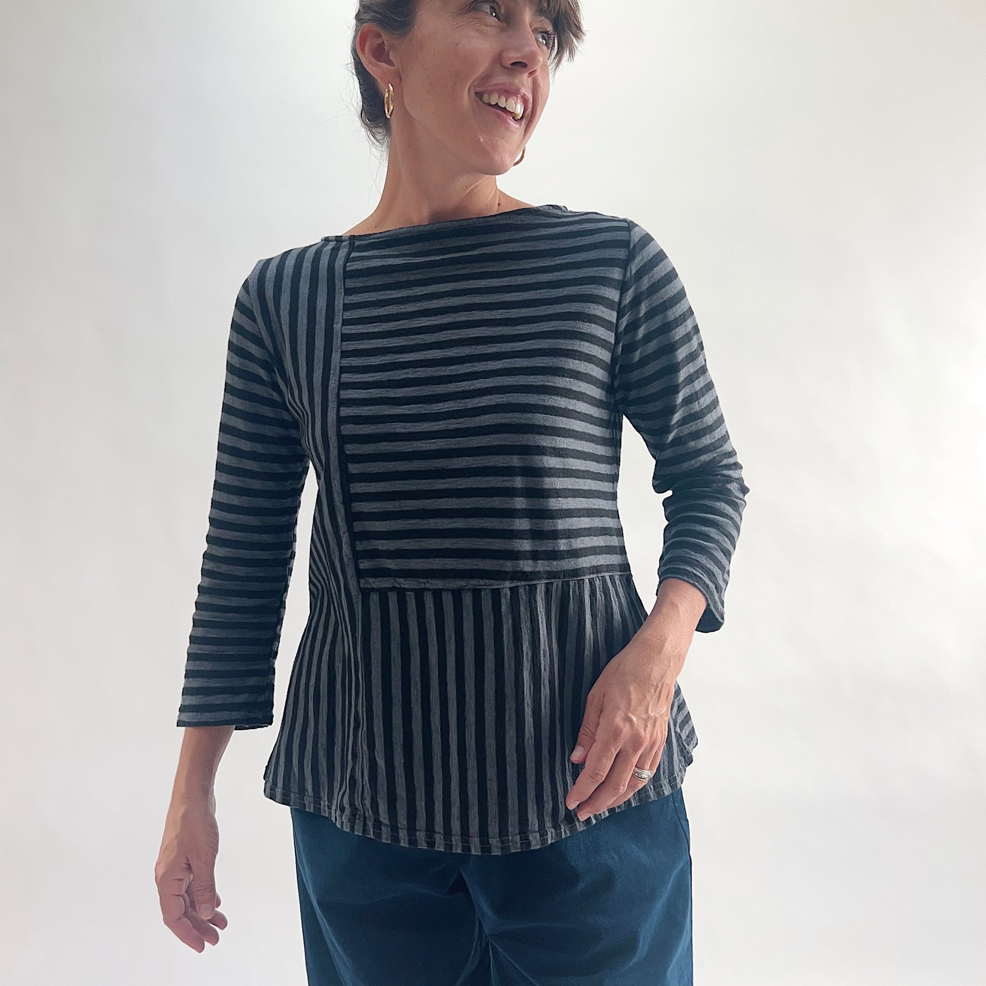 Smiling front view of model wearing a black and white patchwork stripe A-line boatneck top, showing the mixed horizontal and vertical stripe panel construction and flared A-line silhouette.
