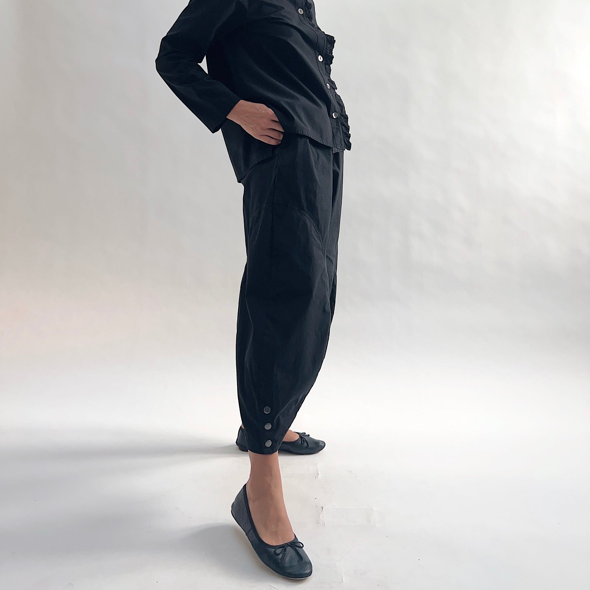 Close front view of model wearing Eleven Stitch black snap doreen pant with hands in pockets, showing elastic waistband, wide balloon leg, side seam pockets, snap buttons at ankle hem, and black color, styled with matching black blossom collar shirt and black ballet flats