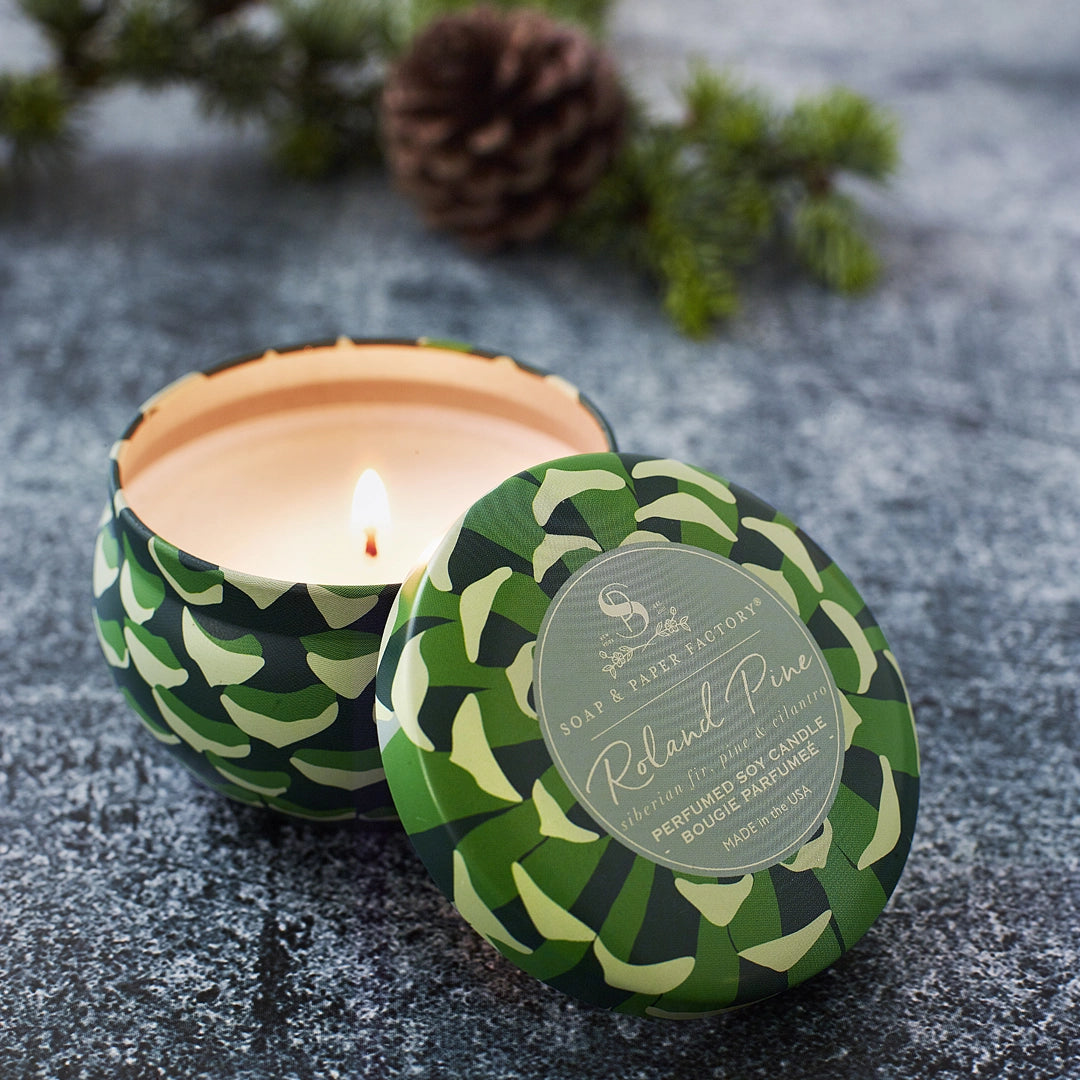 Lit Roland Pine small soy candle and labeled green patterned lid on a textured gray surface, with a pinecone behind.
